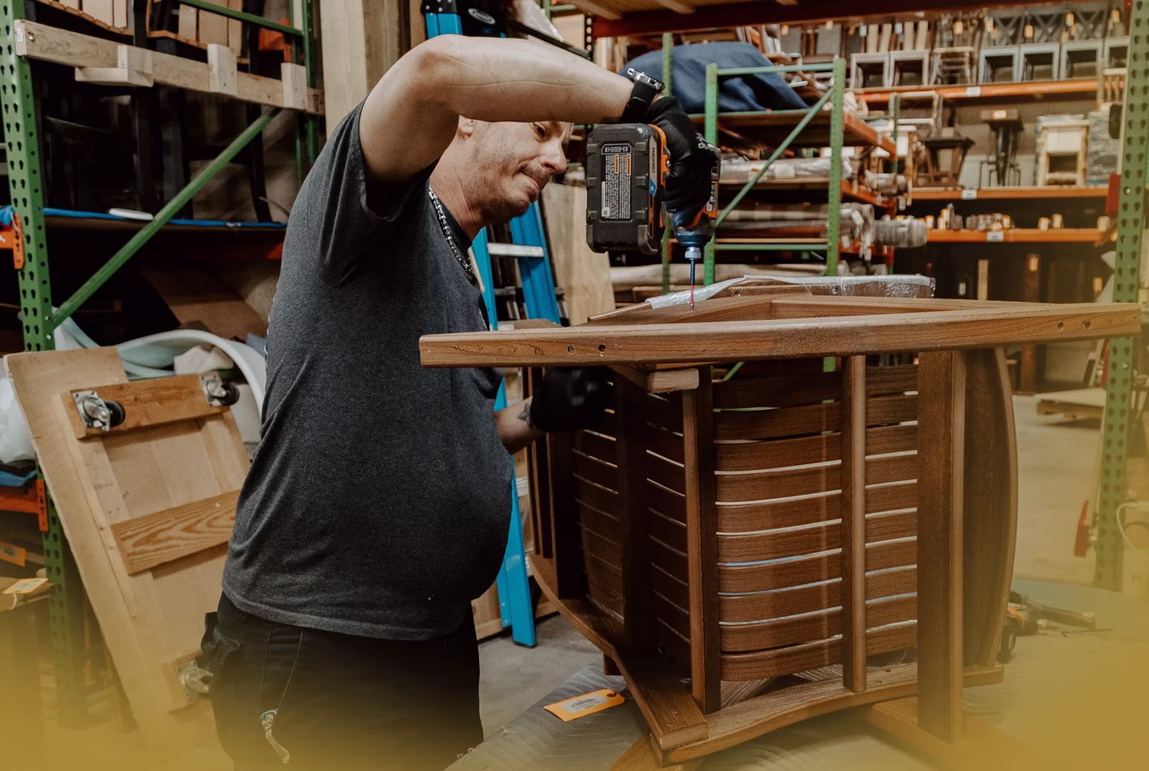 new (257 of 396) A man uses a power drill on a wooden structure in a workshop with shelving in the background.