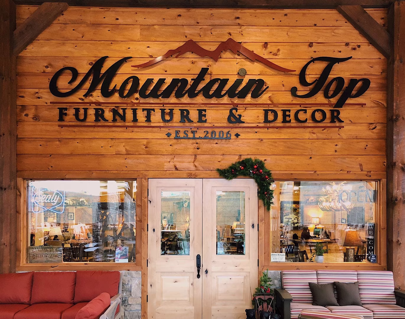 The rustic wooden storefront of Mountain Top Furniture & Decor, featuring white double doors, large display windows, and outdoor seating with red and striped sofas.