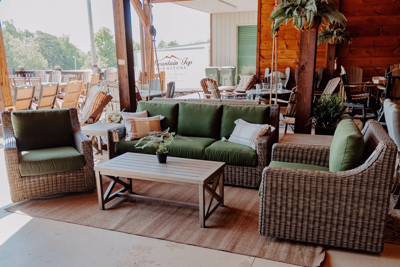 A wooden patio with a green sofa, armchairs, and coffee table is covered by a wooden pergola, accompanied by tables, chairs, and a tree, all set against a brick wall and white building.