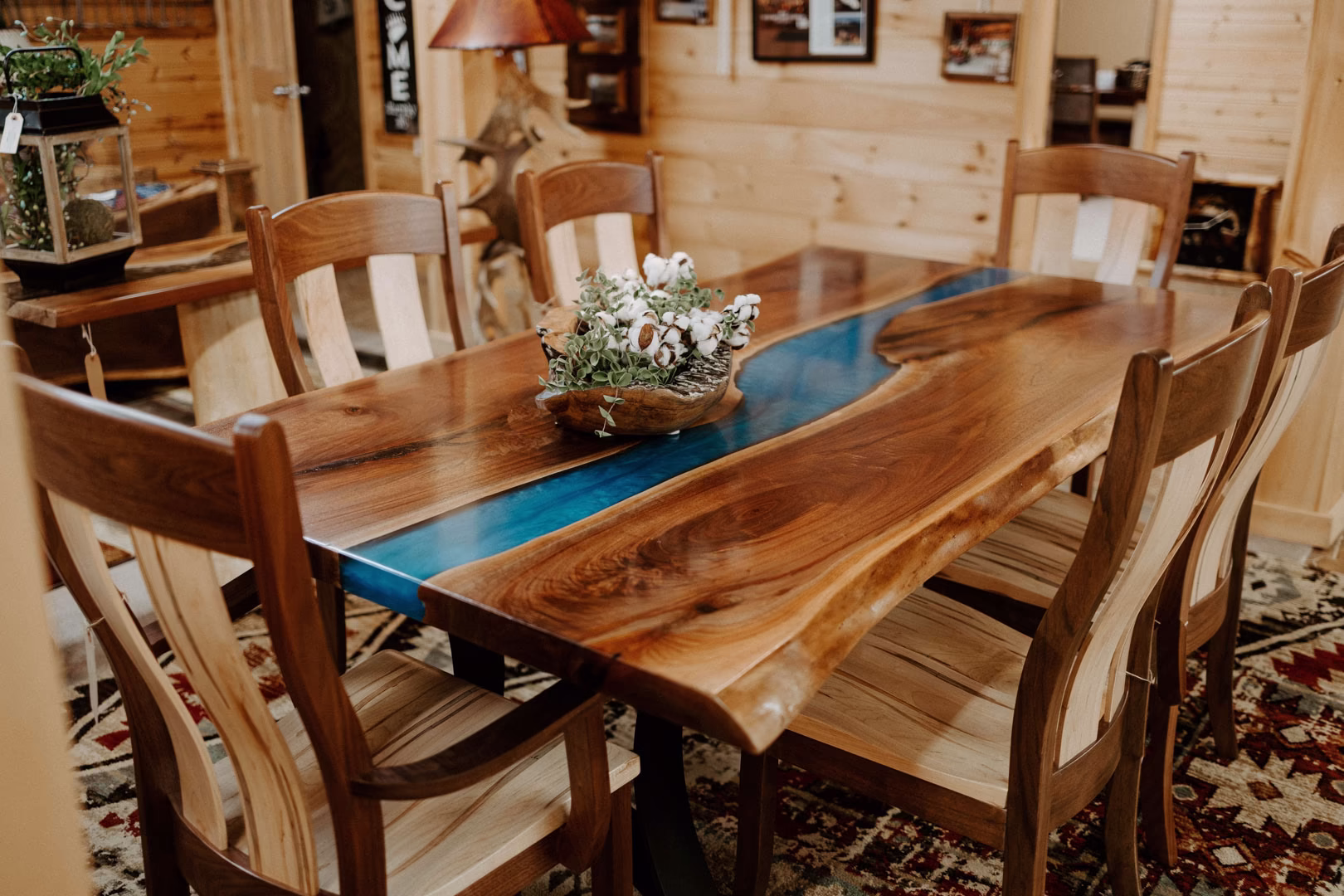 A wooden table with a blue epoxy river running through it, a vase of flowers, and wooden chairs is set against a red and black patterned rug in a cozy room.