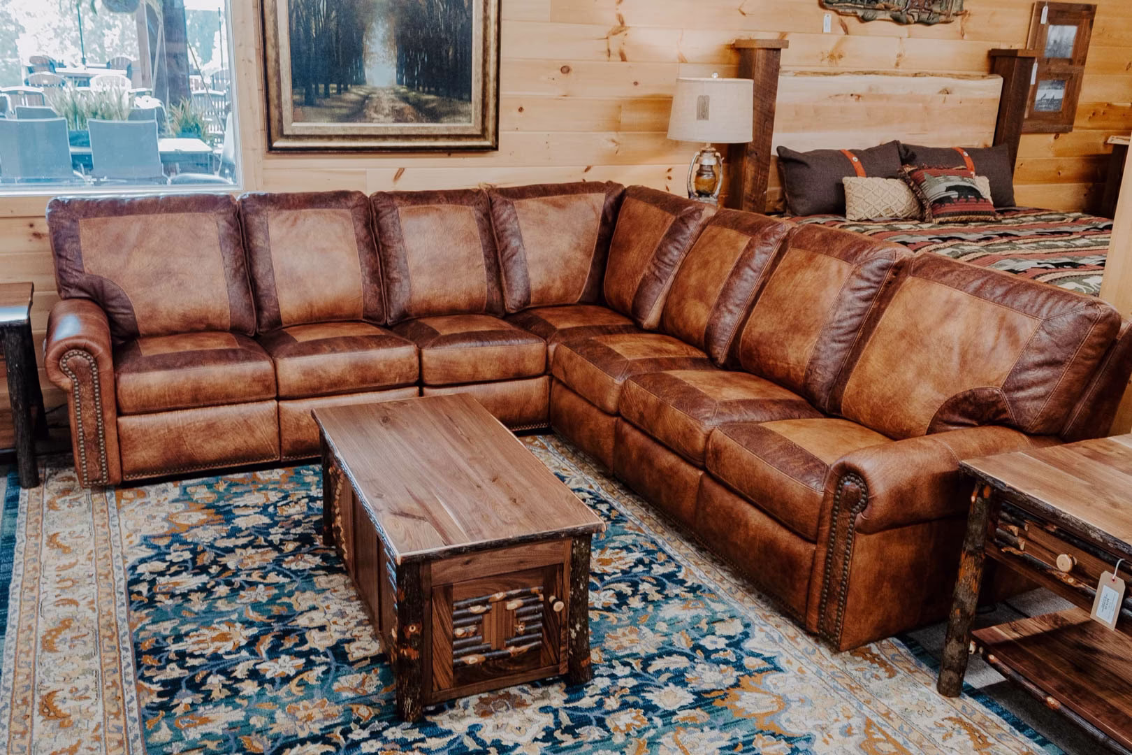 A brown leather sectional sofa with wooden coffee table and blue rug is accompanied by a wooden nightstand and a painting of a forest, in a cozy living room with light wooden walls.