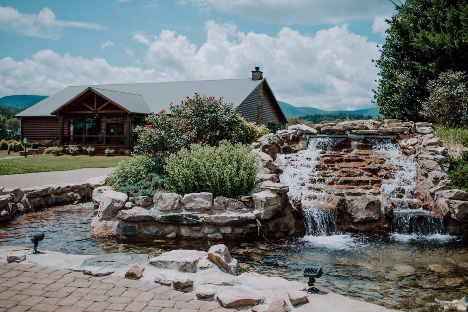 new (277 of 396) A two-story wooden building with a brown roof and chimney, accompanied by a stone fountain and a pond, is nestled within a lush natural setting.