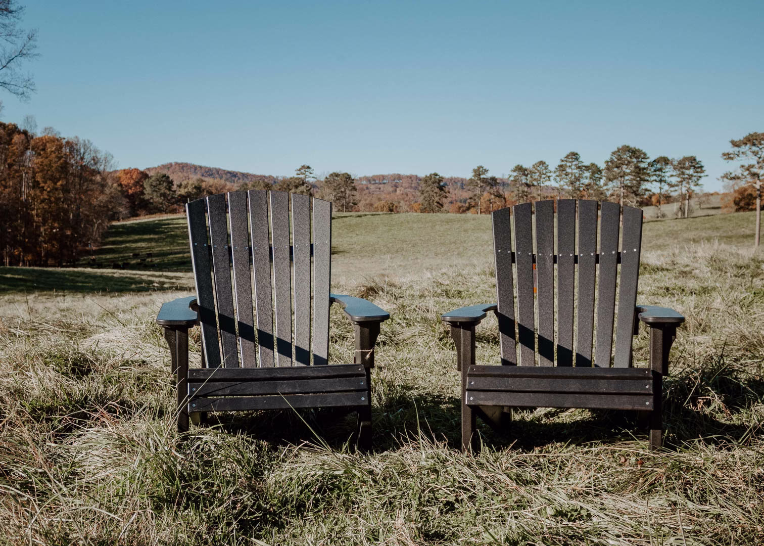 Two black Adirondack chairs face each other on a grassy hill, backed by a clear blue sky, trees, and a distant mountain range.