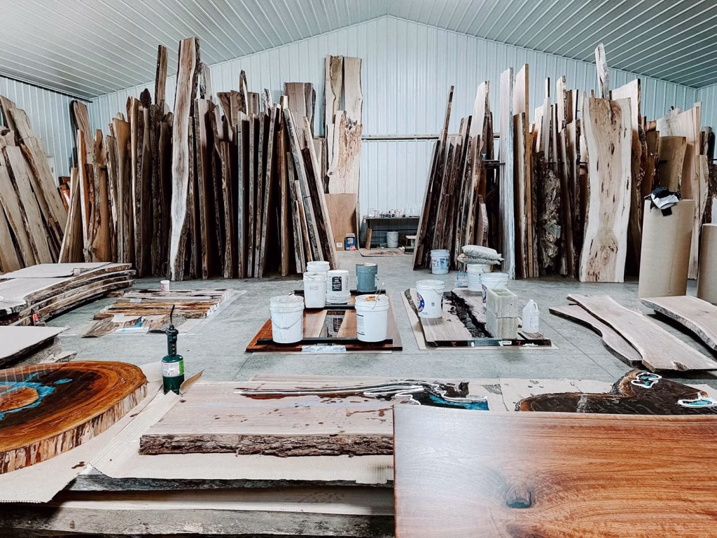 A bustling workshop filled with various sizes and colors of wooden slabs, stacks of lumber, and paint cans, features a natural light source and tools for woodworking.
