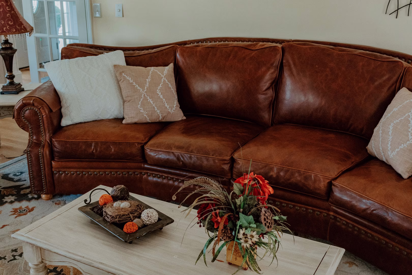new (60 of 396) A cozy living room features a brown leather couch with white and beige throw pillows, a white coffee table with a black tray and a red and white vase, and a red lamp with a gold base.