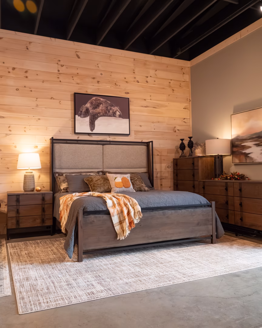 new (77 of 396) A wooden bed with a gray headboard and orange and yellow throw blankets is accompanied by a wooden dresser, nightstands, and a large framed picture of a brown bear, set against a wooden wall with exposed beams.