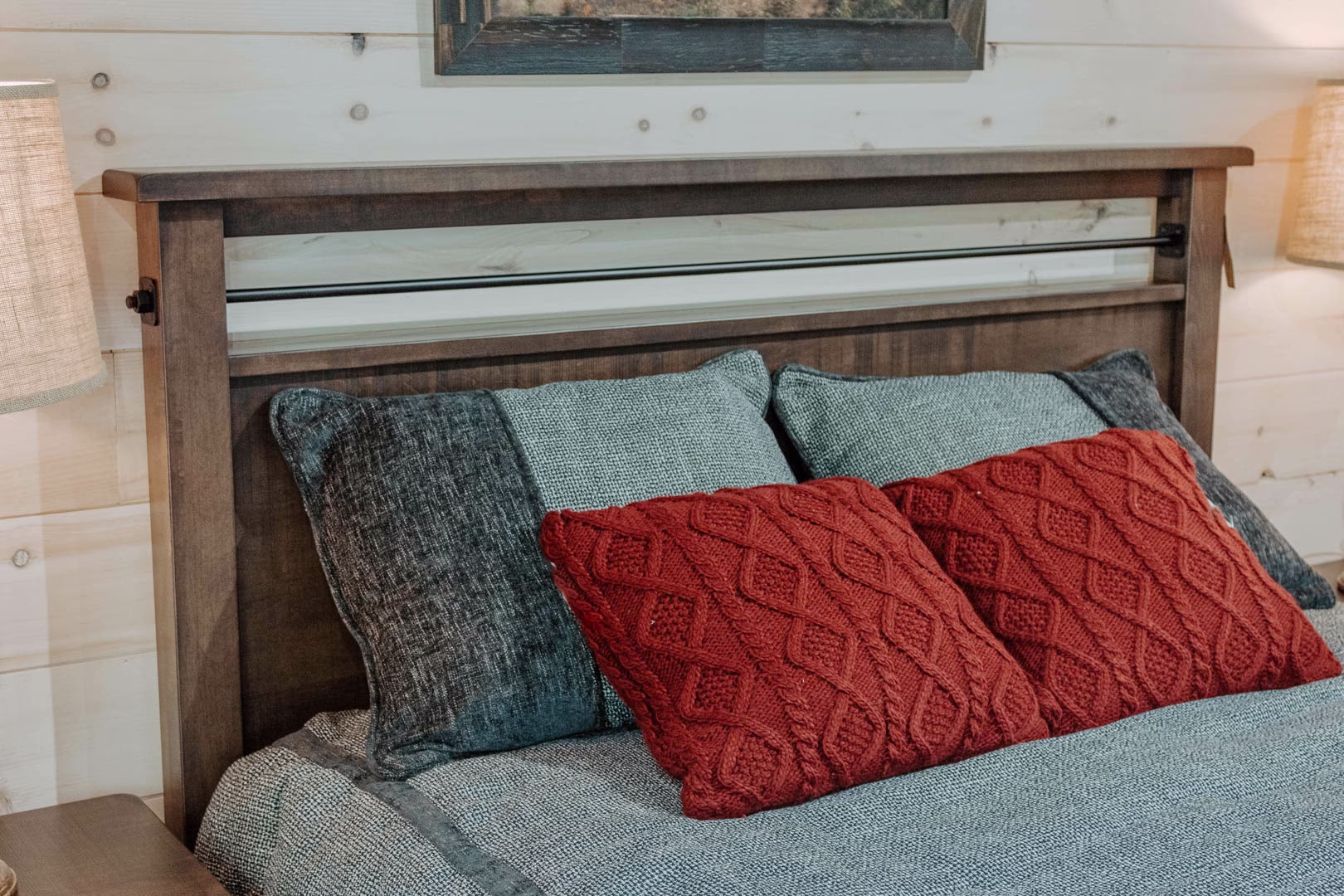 new (81 of 396) A wooden bed with a headboard and footboard, flanked by gray and red throw pillows, is set against a white wooden wall with a black picture frame and lamp.