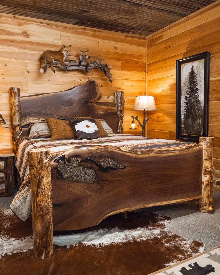 A rustic bedroom features a live-edge wooden bed with white and brown pillows, a deer head above, and a lamp on a nearby nightstand.