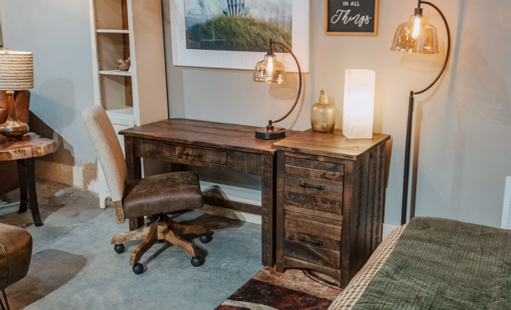 barn-floor-writing-desk-1 A wooden desk with a lamp, vase, and picture sits against a white wall, accompanied by a wooden chair and a green rug, illuminated by natural light.