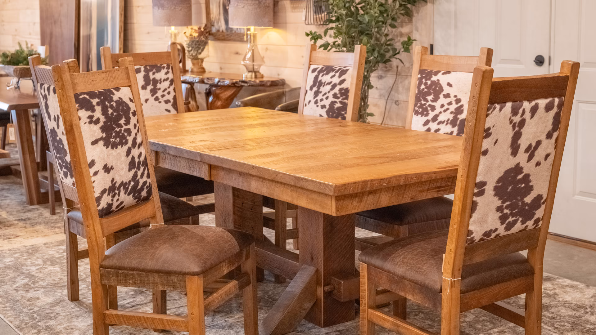 A rustic wooden dining table with six cowhide-patterned chairs is set for a meal in a warm, inviting room with wooden walls, a hanging plant, and a vase of flowers.