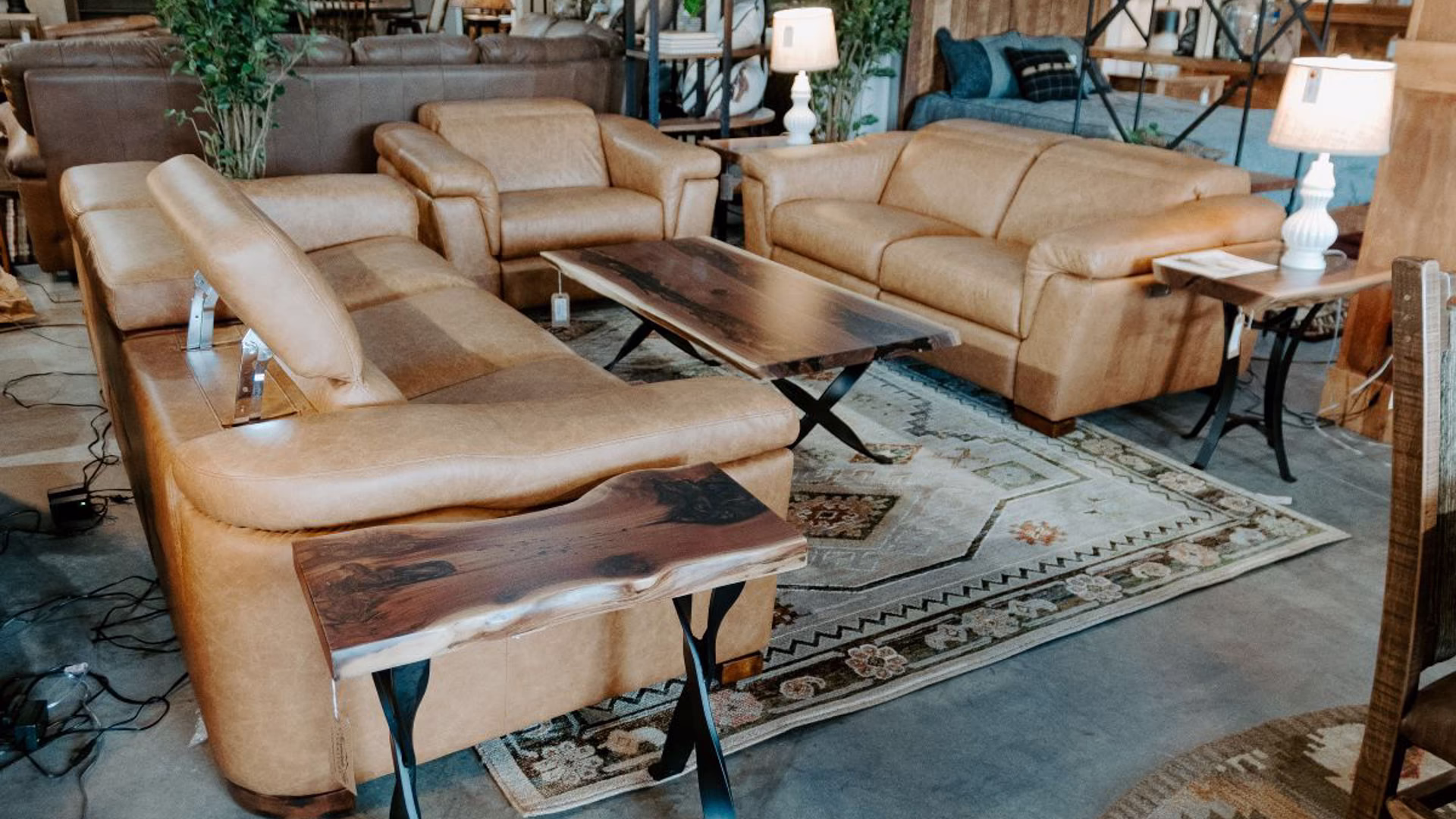 bergamo-ponza-collection-featured-image A furniture showroom features a brown leather sofa, loveseat, and armchairs arranged around a wooden coffee table, with a rug, bookshelf, and plants adding to the cozy ambiance.
