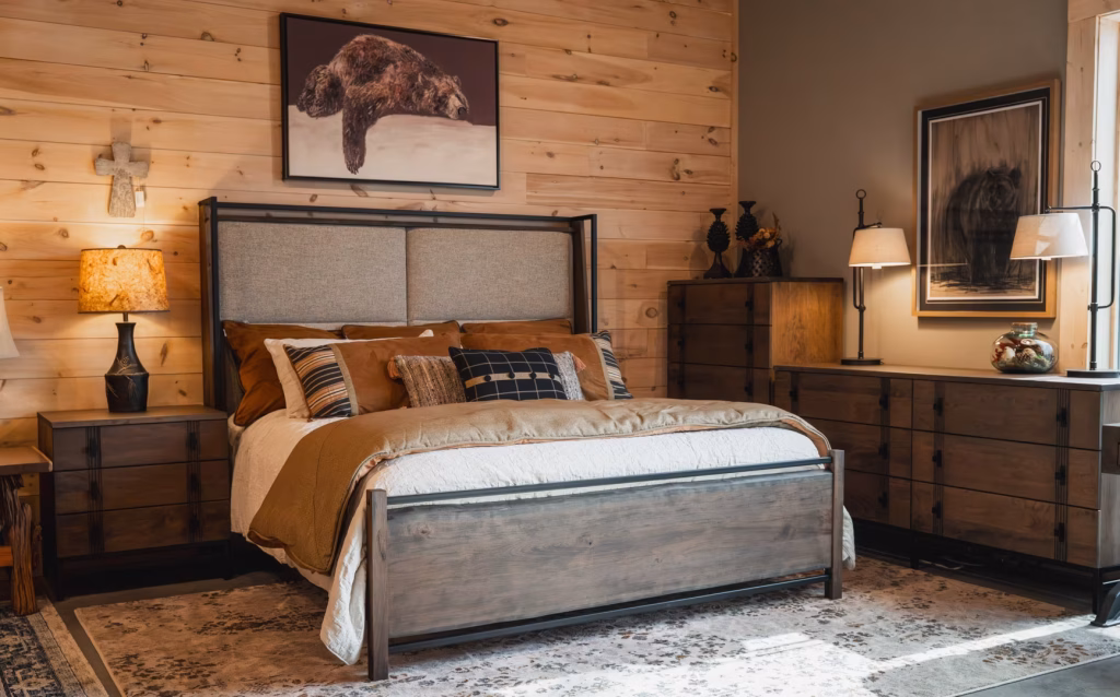 berkshire-collection-featured-image A wooden bed with brown comforter and pillows, a wooden nightstand, a dresser, and a black lamp create a warm, rustic bedroom with a mountain-themed artwork.