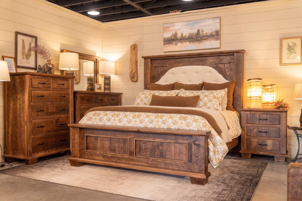 blaire-collection-featured-image A bedroom display features a wooden bed with gold and white comforter, flanked by wooden nightstands and lamps, with framed pictures and paintings on the white-painted walls.