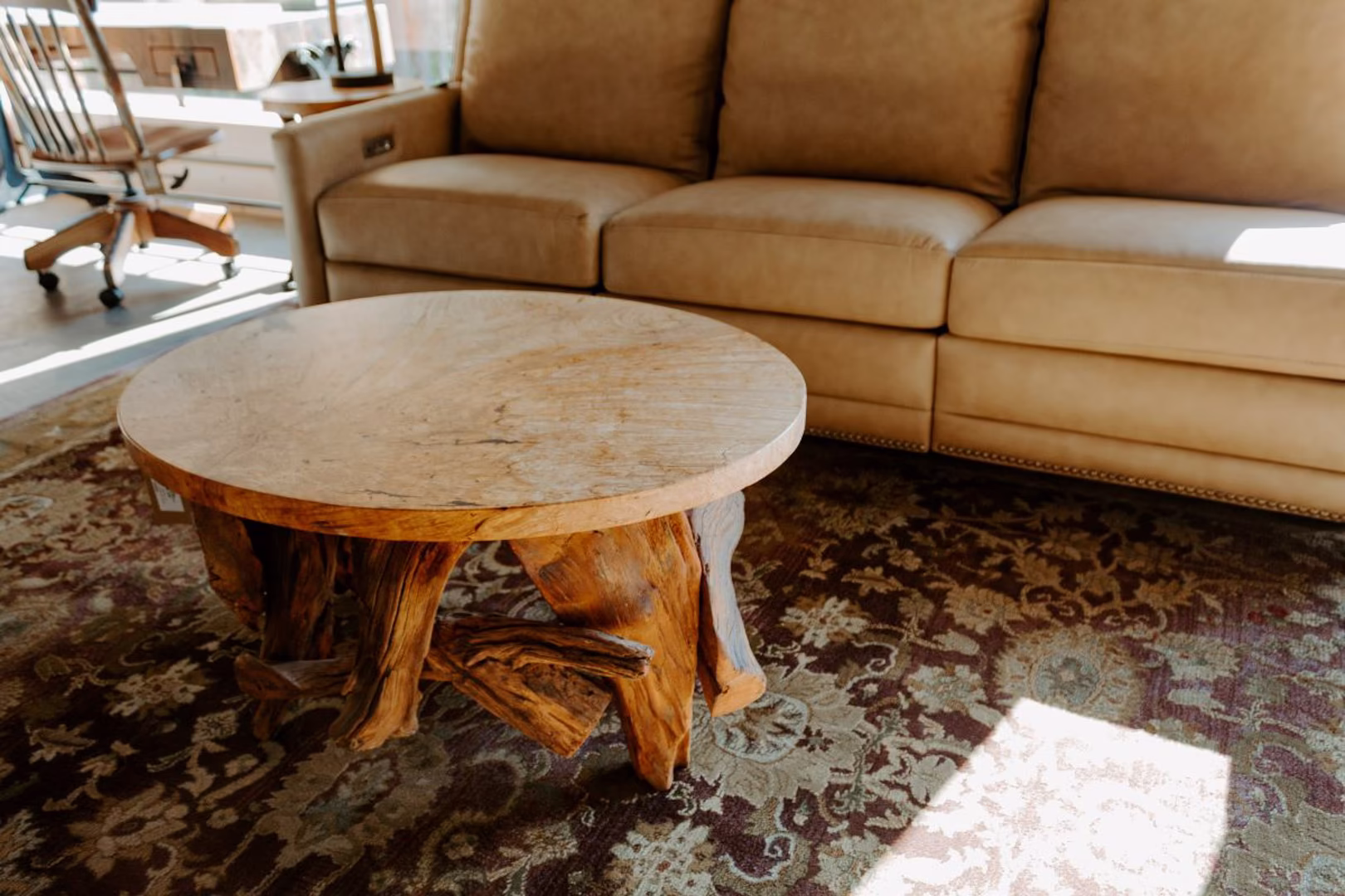 A round wooden coffee table with a natural wood top and branch legs sits on a vibrant floral rug, accompanied by a beige sofa and a wooden rocking chair.