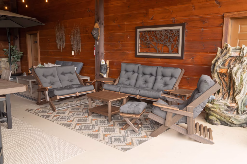 A cozy outdoor patio features a gray sofa, wooden coffee table, and recliner, with earth-toned decor and a tree trunk sculpture, enclosed by a wooden fence.