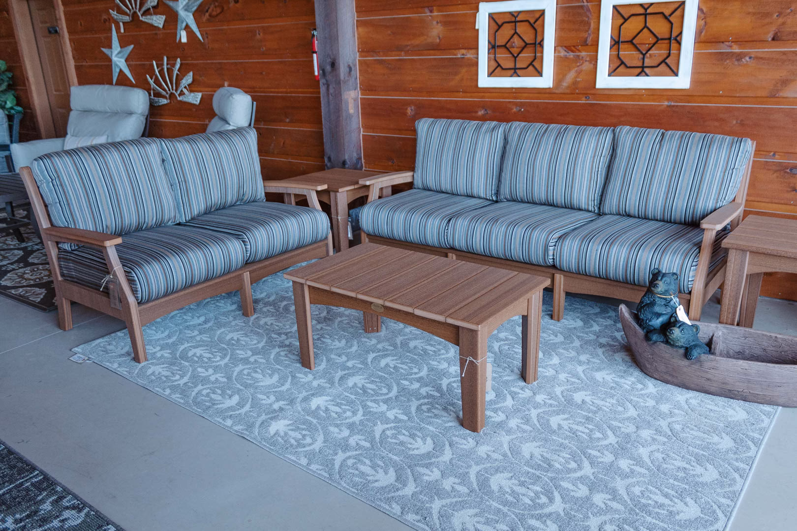 classic-terrace-stone-collection-featured-image An outdoor furniture set, including a blue and white striped couch, white coffee table, and wooden side table, is arranged on a gray rug with a geometric pattern, accompanied by a wooden bench and decorative wall art.