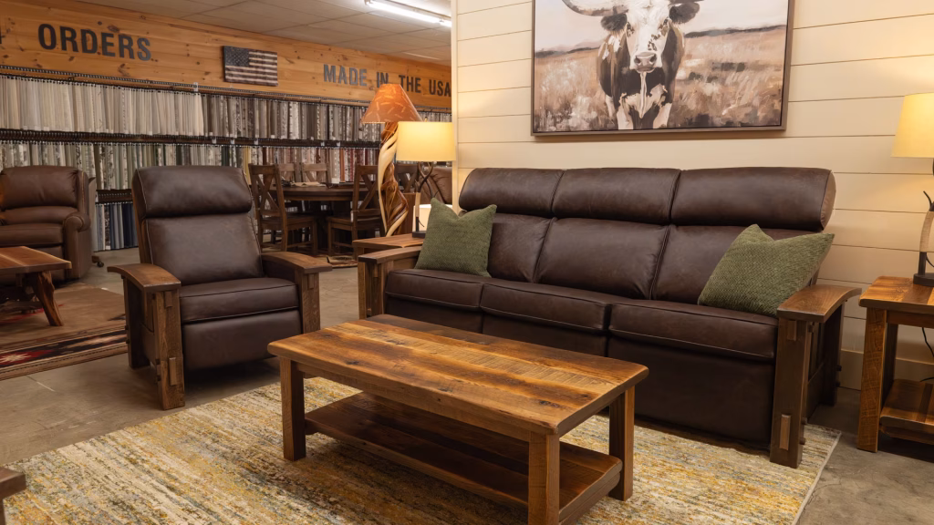 A brown leather recliner and sofa, accompanied by a wooden coffee table and side table, are arranged in a room with a wooden wall, a framed cow picture, and a bookshelf.