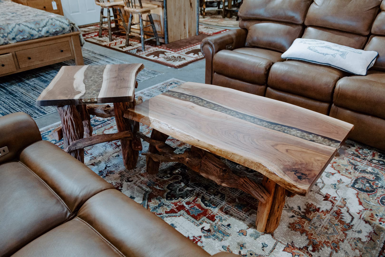 A furniture showroom features a brown leather armchair, wooden coffee table with resin top, and a floral sofa, arranged on a vibrant rug.