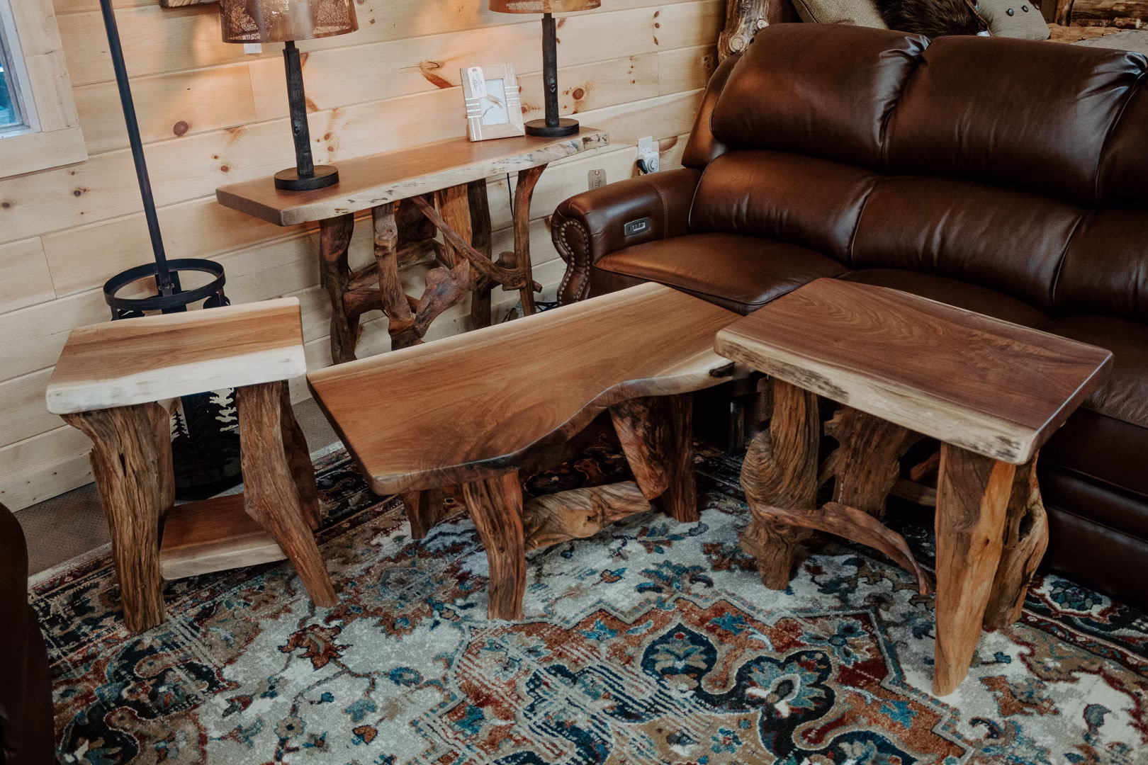 driftwood-walnut-collection-featured-image A cozy living room corner features a wooden coffee table, side tables, a brown leather couch, and a wooden wall, with a rug and lamp adding warmth and charm.