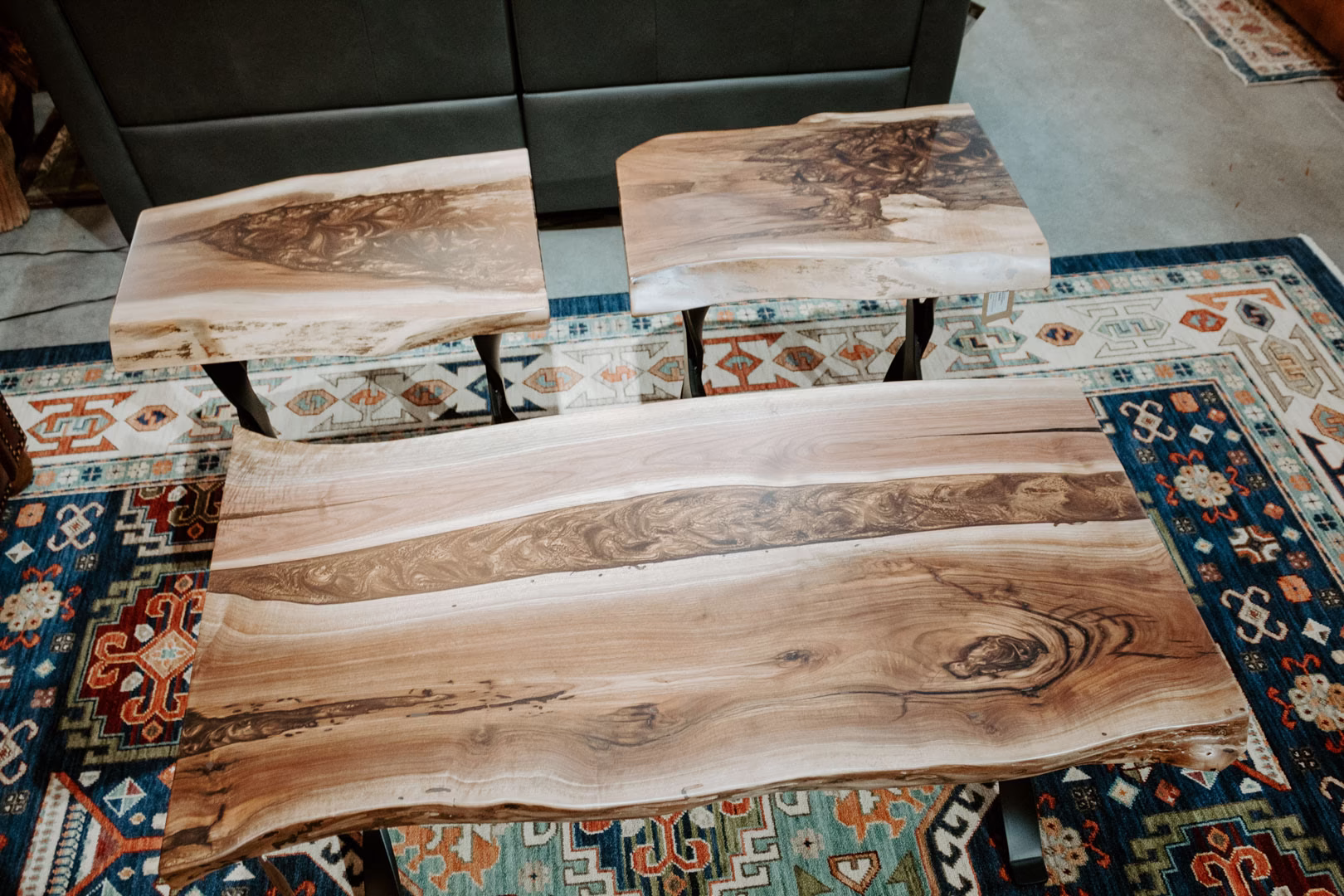 espresso-epoxy-slab-collection-featured-image A light-colored wooden coffee table with a natural edge and rich grain sits atop a vibrant, intricately patterned rug, with a black leather couch nearby.