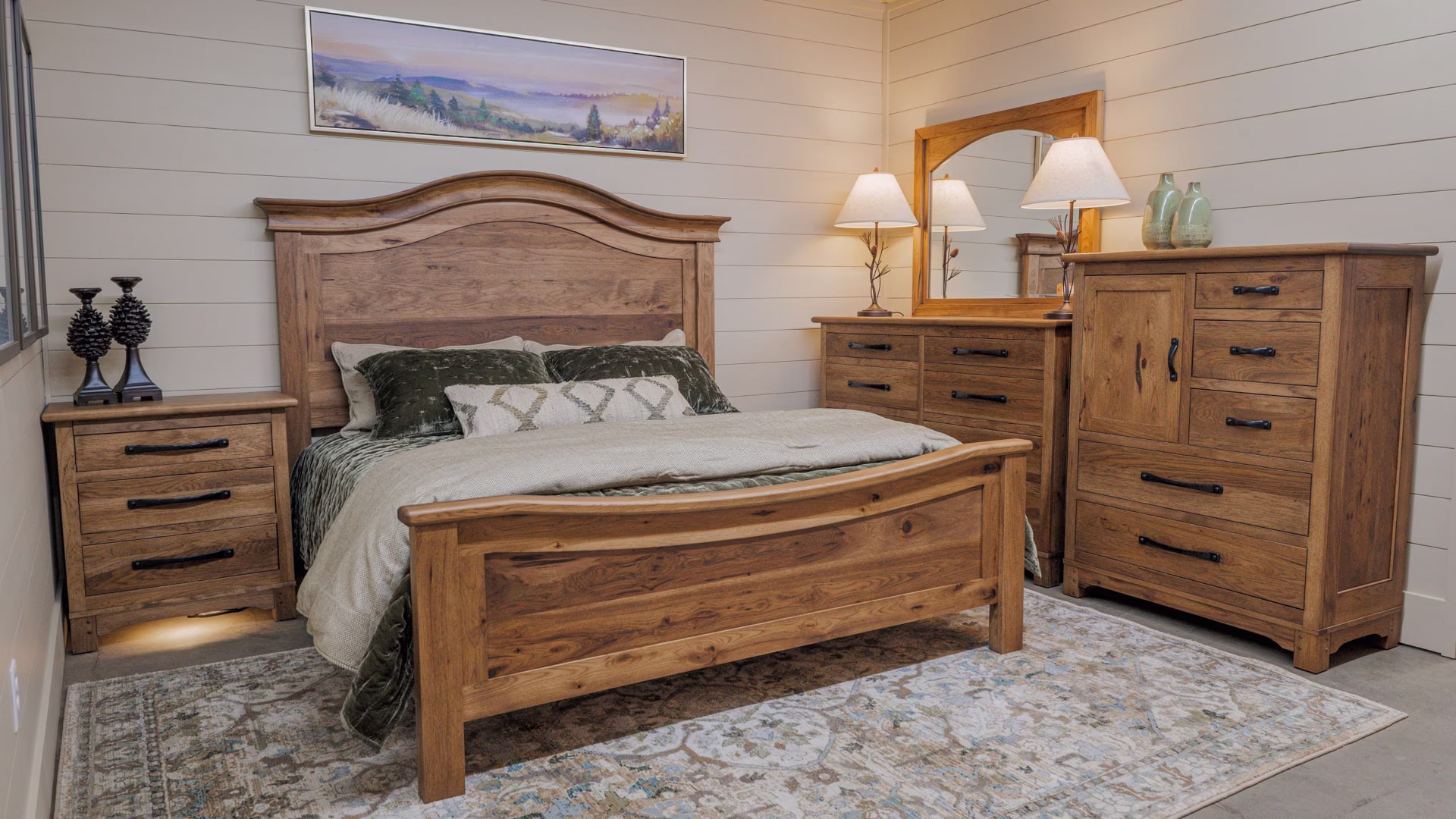 A wooden bed with a white and gray striped comforter, accompanied by a wooden nightstand, dresser, and lamp, occupies a cozy bedroom with a landscape painting and beige carpet.