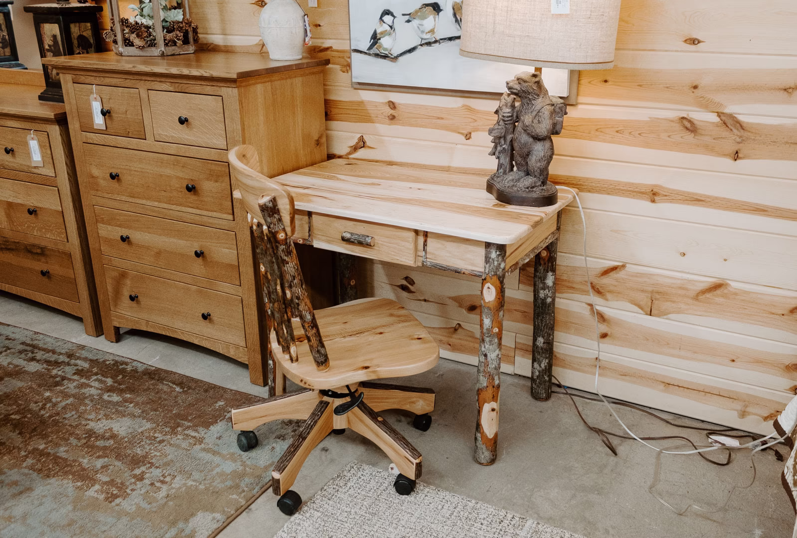 A wooden desk with a lamp and bear statue, accompanied by a wooden dresser and plant, occupies a cozy corner in a room with wooden walls.