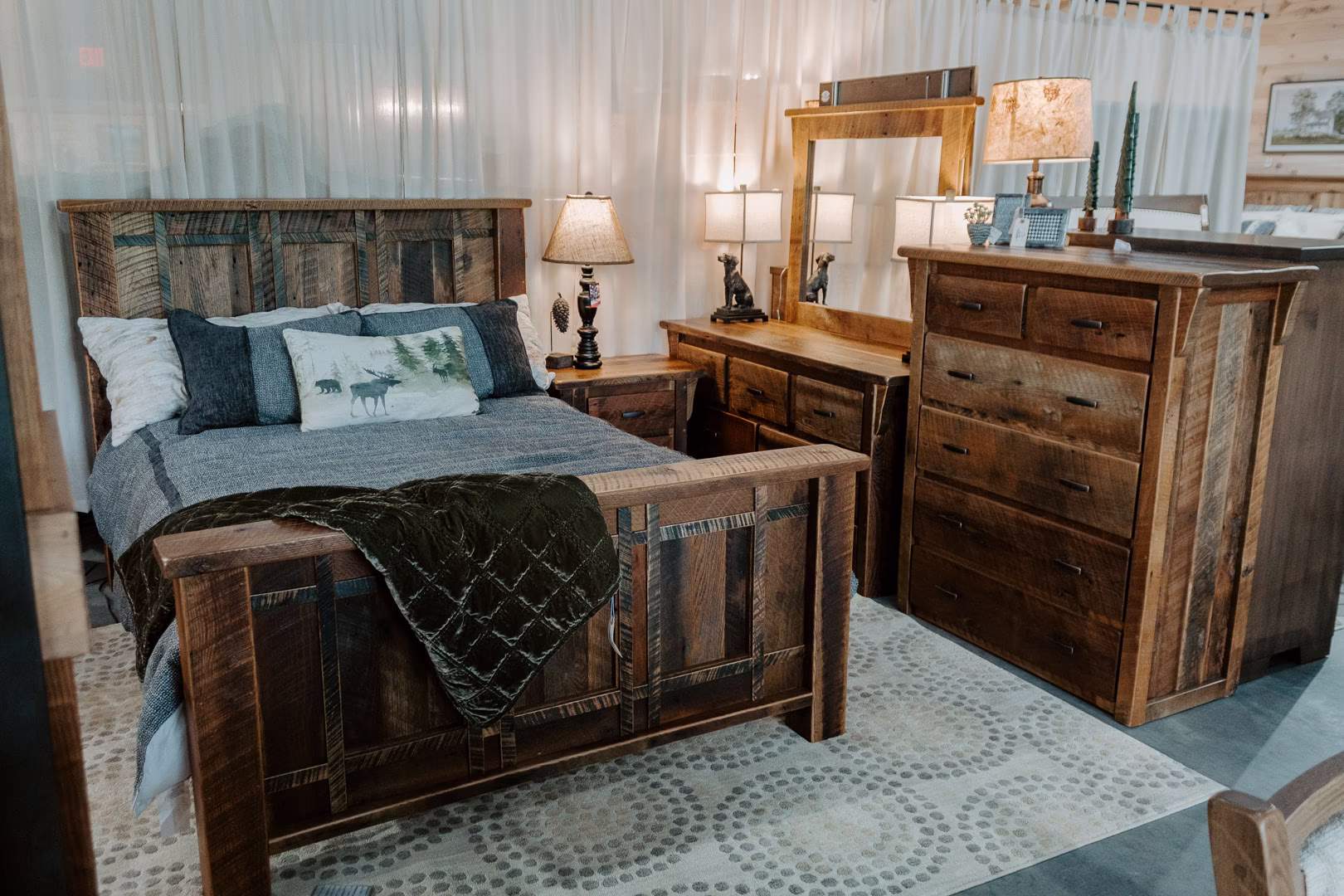 A bedroom display showcases a rustic wooden bed with gray comforter, dark green throw, and white and gray pillows, accompanied by a wooden dresser, lamp, and nightstand.