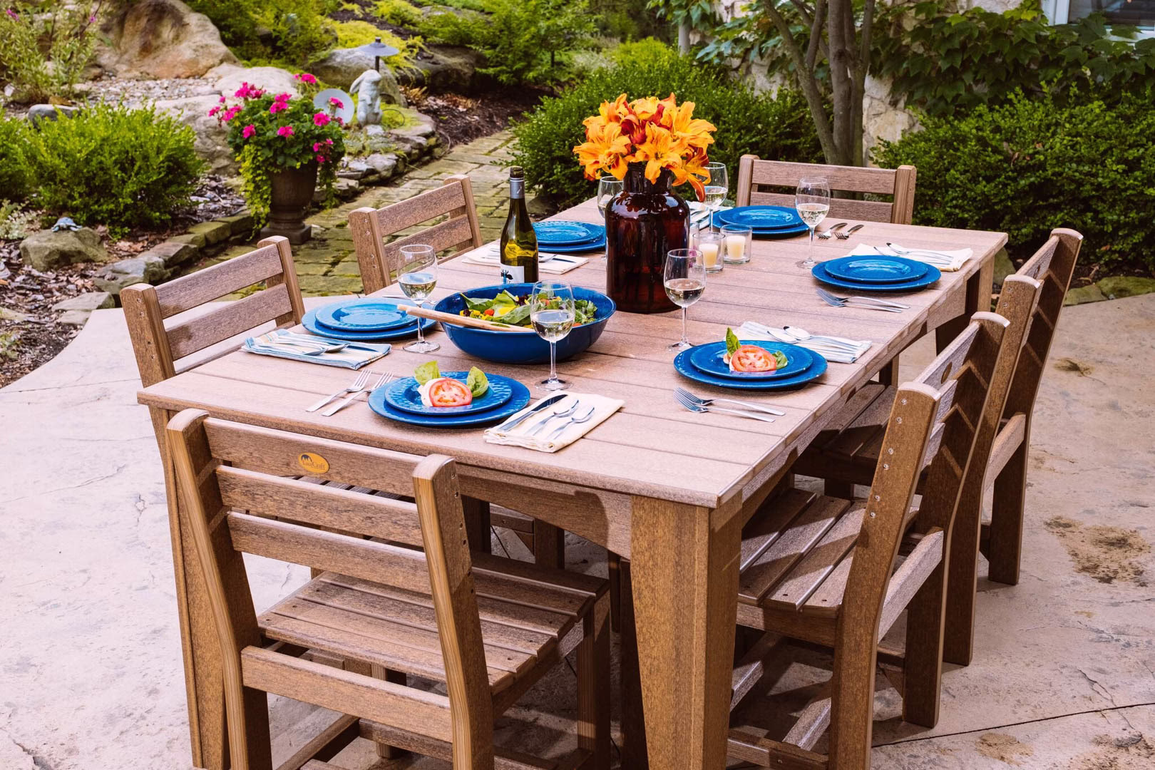 island-dining-collection-featured-image An outdoor dining setup features a rectangular wooden table with blue plates, silverware, and a vase of orange and yellow flowers, set on a patio with natural lighting and a garden backdrop.