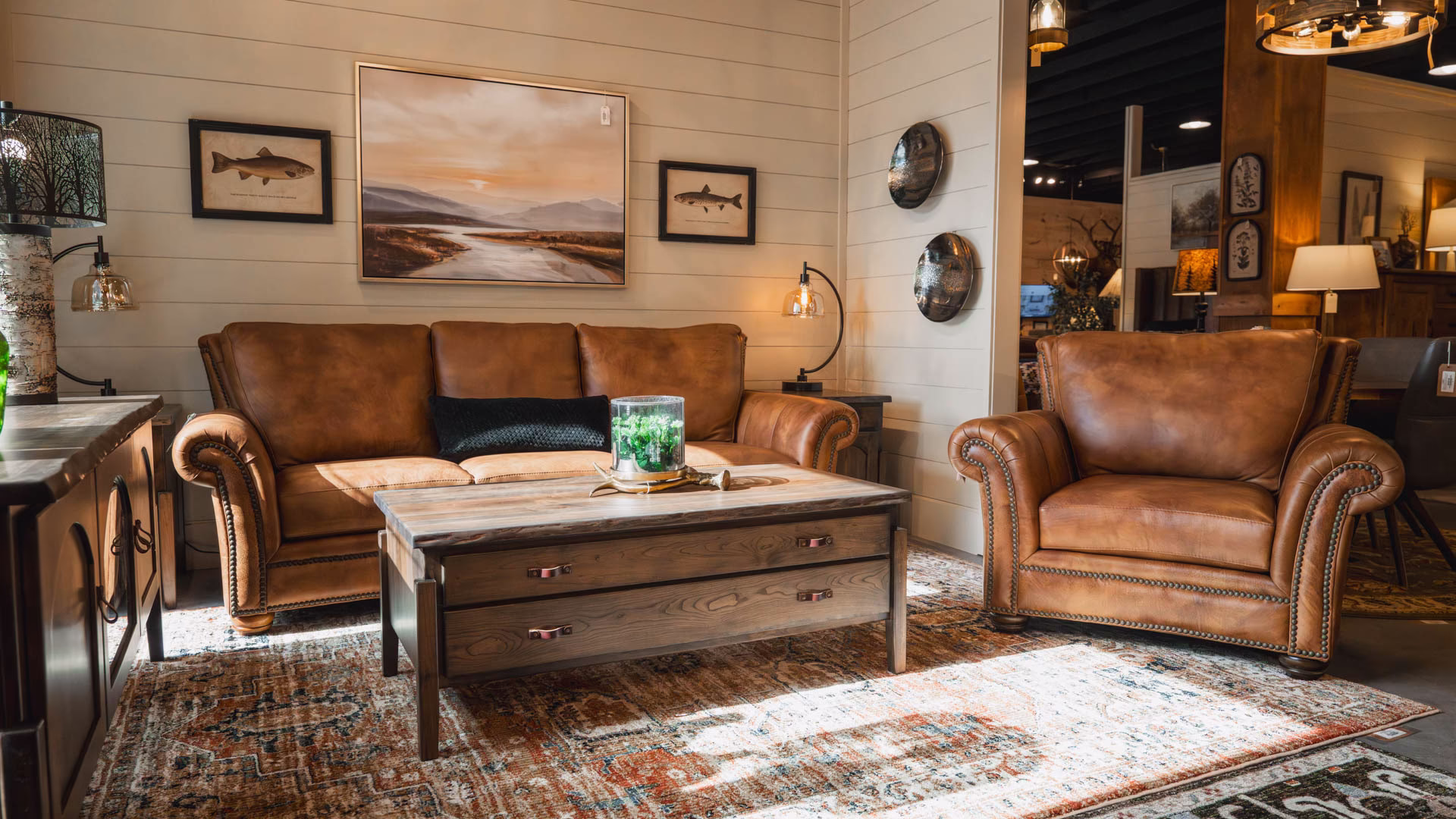A brown leather sofa and armchair, accompanied by a wooden coffee table and green plant, occupy a cozy living room with a brick wall, wooden floor, and framed paintings.