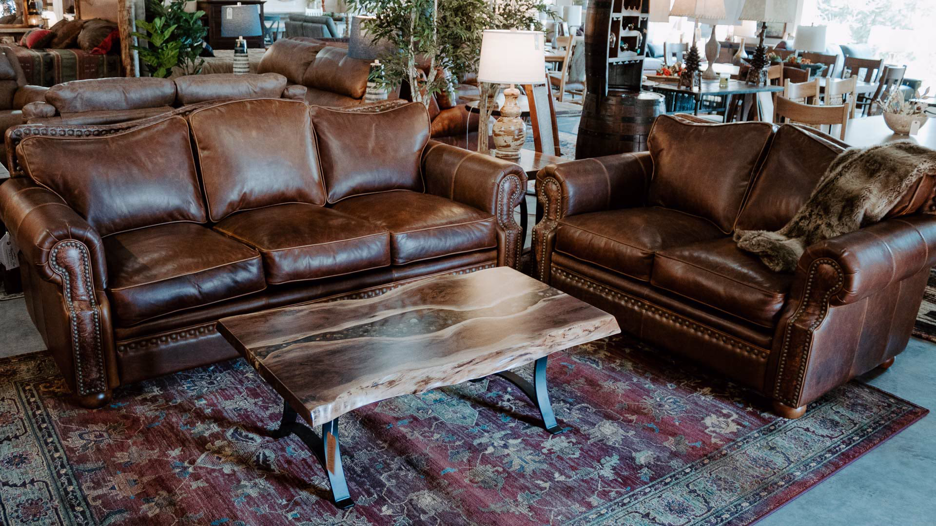 A brown leather sofa and loveseat, with a wooden coffee table and black metal base, occupy a cozy living room with a red and blue rug, white lamp, and green plant.