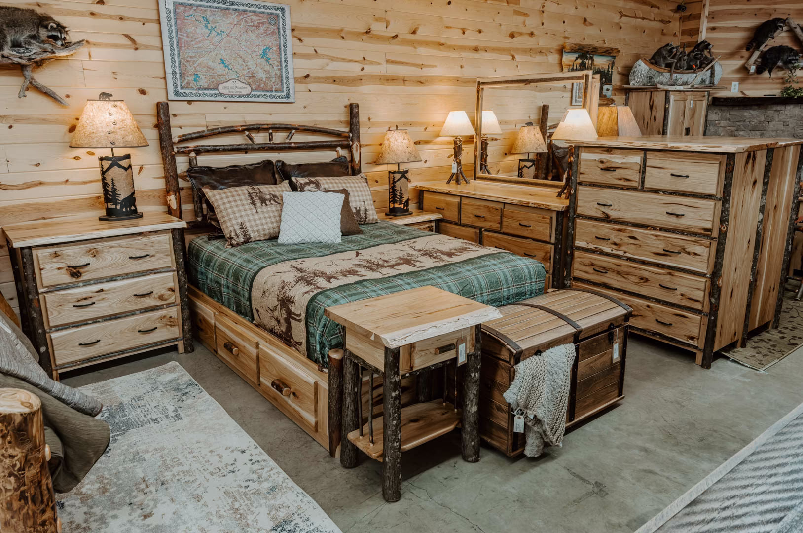 A rustic bedroom setting features a wooden bed with a green and brown plaid comforter, a wooden dresser, a trunk bed, and a framed map, all complemented by lamps and a rug.