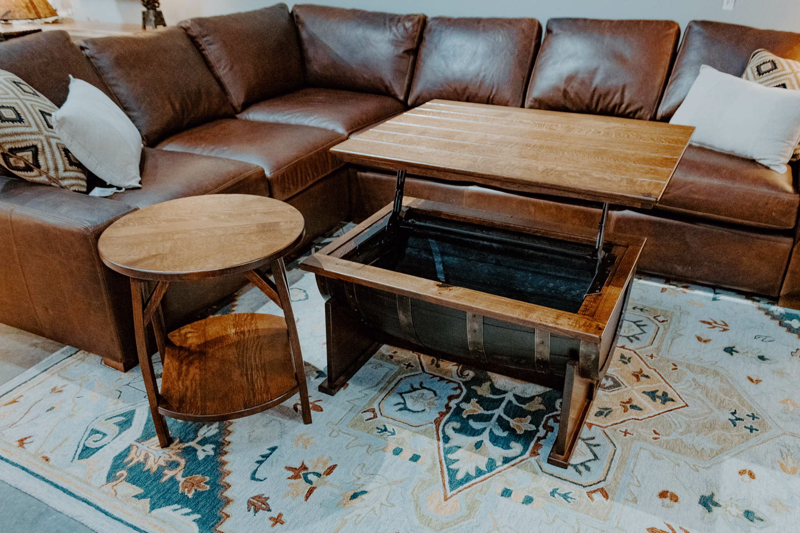 lift-top-barrel-collection-featured-image A brown leather sofa with throw pillows sits on a patterned rug, accompanied by a wooden coffee table with a built-in barrel and a wooden side table.
