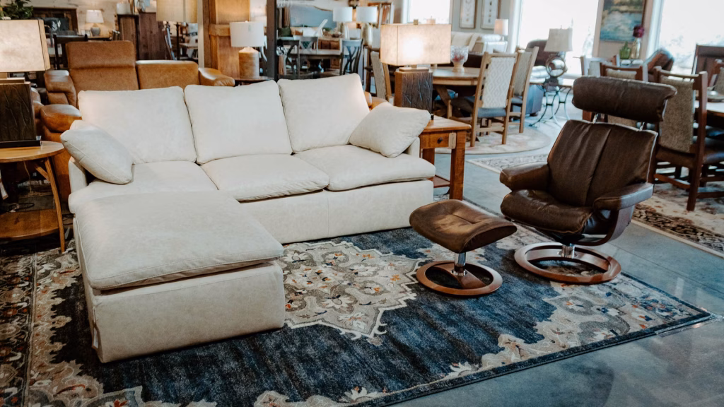 A spacious living room showcases a white sofa, blue and white rug, brown leather armchair, and wooden side table, bathed in natural light.