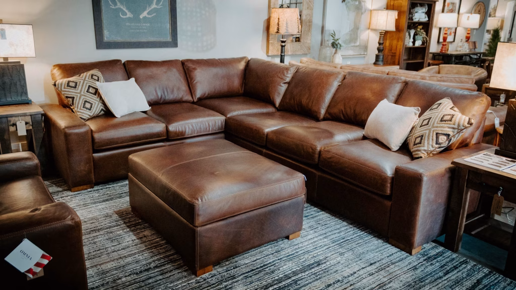 A brown leather sectional sofa with throw pillows and a ottoman, accompanied by a wooden coffee table and lamp, occupies a cozy living room with neutral-toned walls and wooden floors.