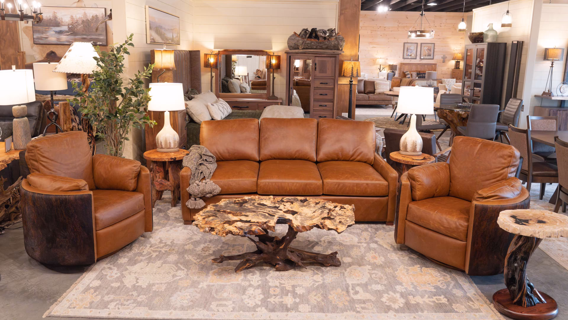A brown leather sofa, two matching armchairs, and a wooden coffee table with driftwood base occupy a cozy living room, accompanied by a side table, lamp, plant, and paintings.