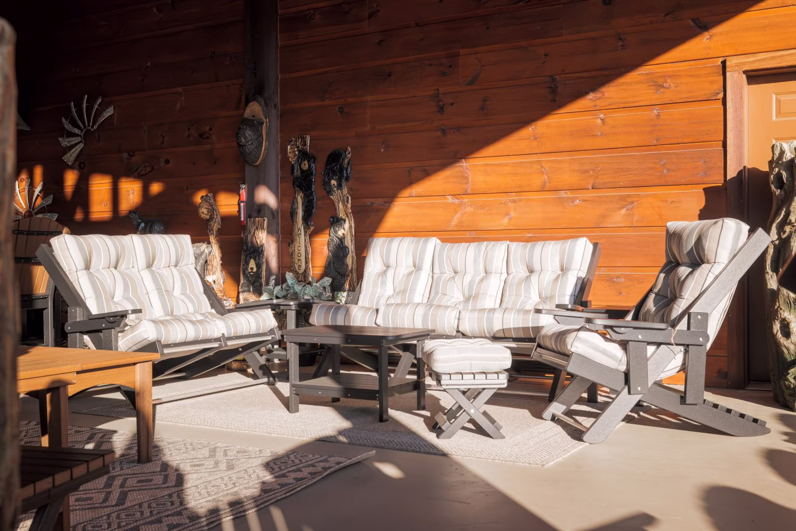 A cozy outdoor patio features a wooden wall with carved trees, a bench with striped cushions, and a table with two chairs, all illuminated by natural light.