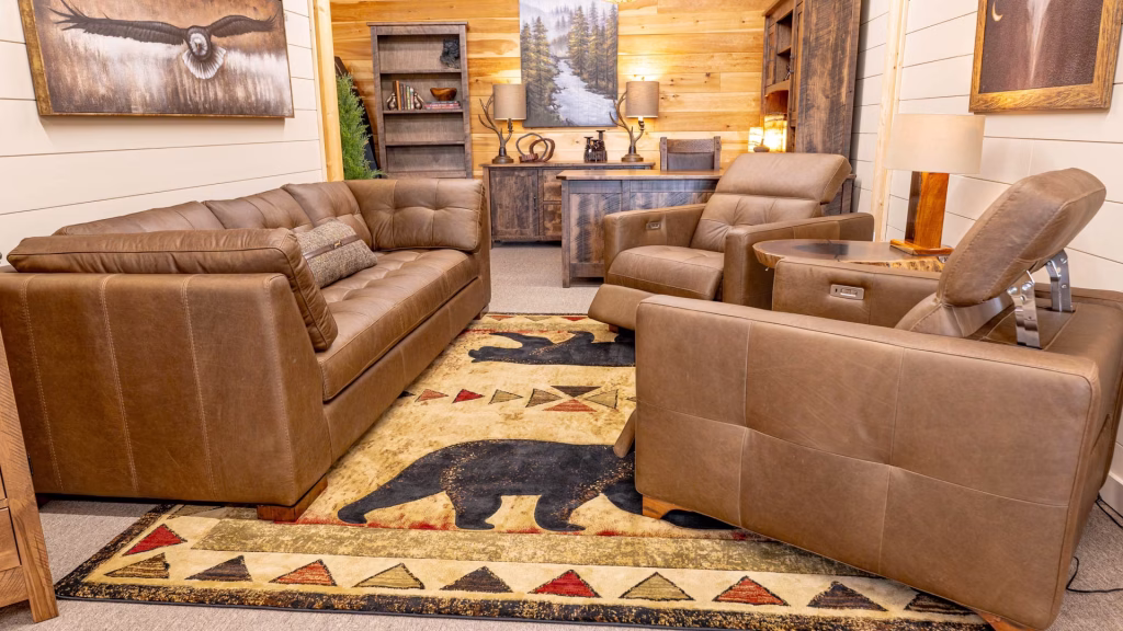 A cozy living room features a brown leather sofa, armchairs, and a wooden coffee table, with a black bear rug, bookshelf, and lamp adding to the rustic charm.