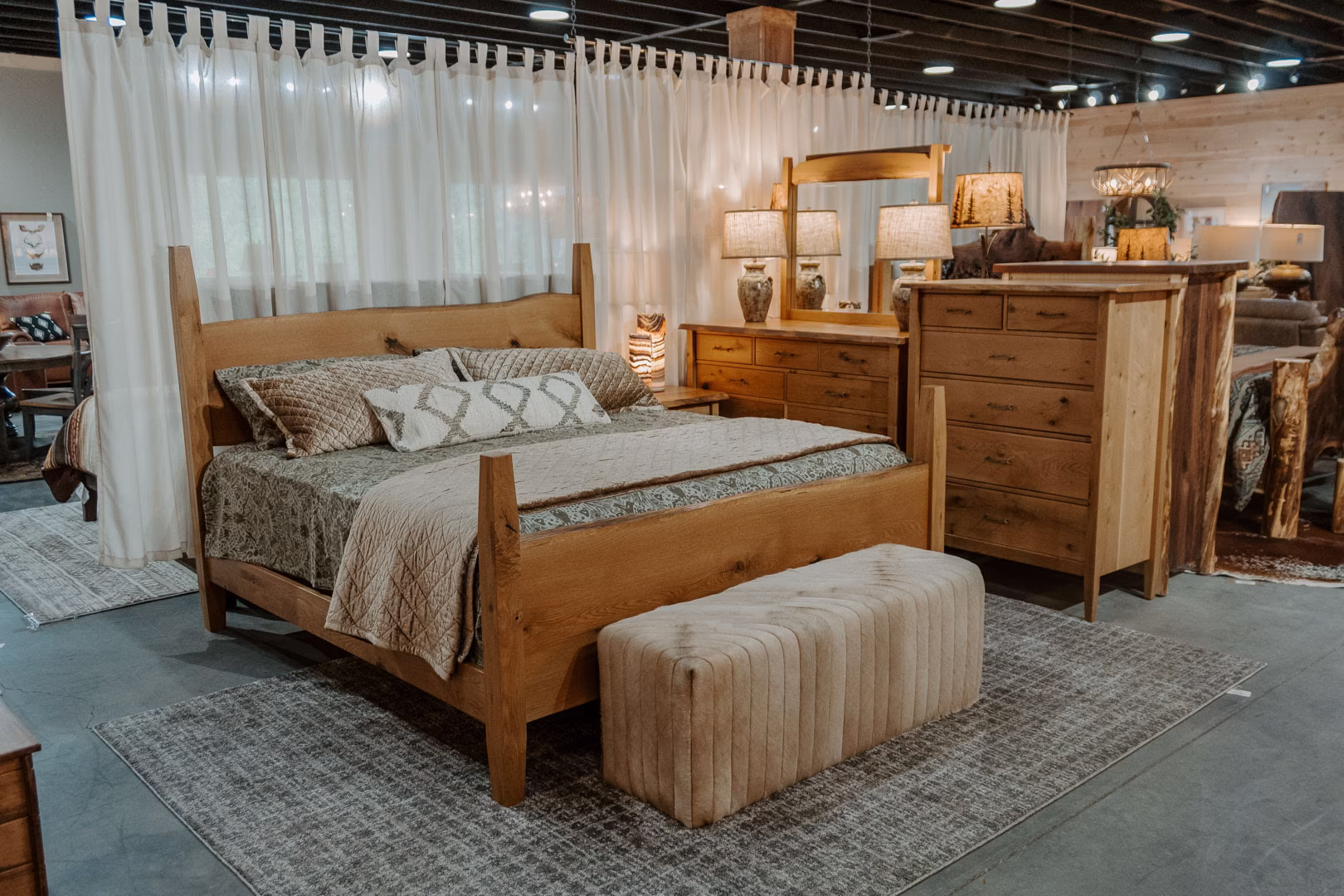 A bedroom display in a furniture store features a wooden bed with gray and white bedding, a wooden dresser, a white armchair, and a gray rug, with white curtains and string lights adding ambiance.