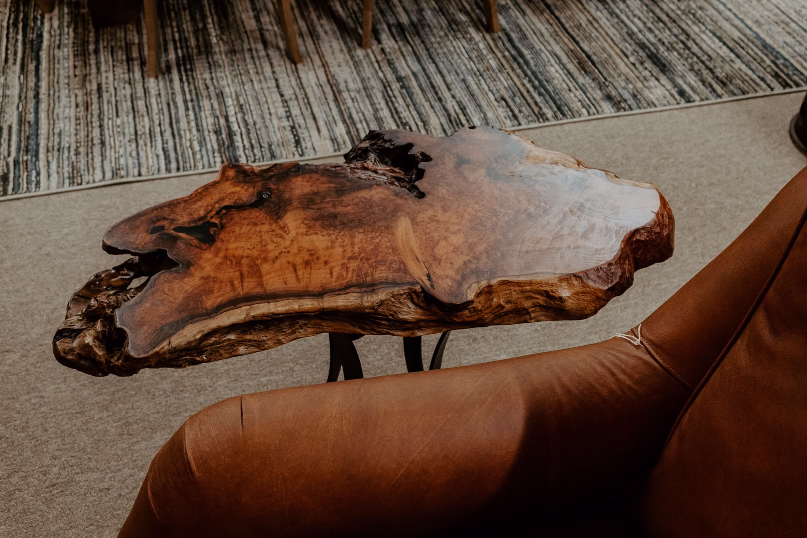 A dark wooden side table with a natural, rough-hewn edge and geometric top sits beside a brown leather armchair, contrasting in the room's neutral color palette.