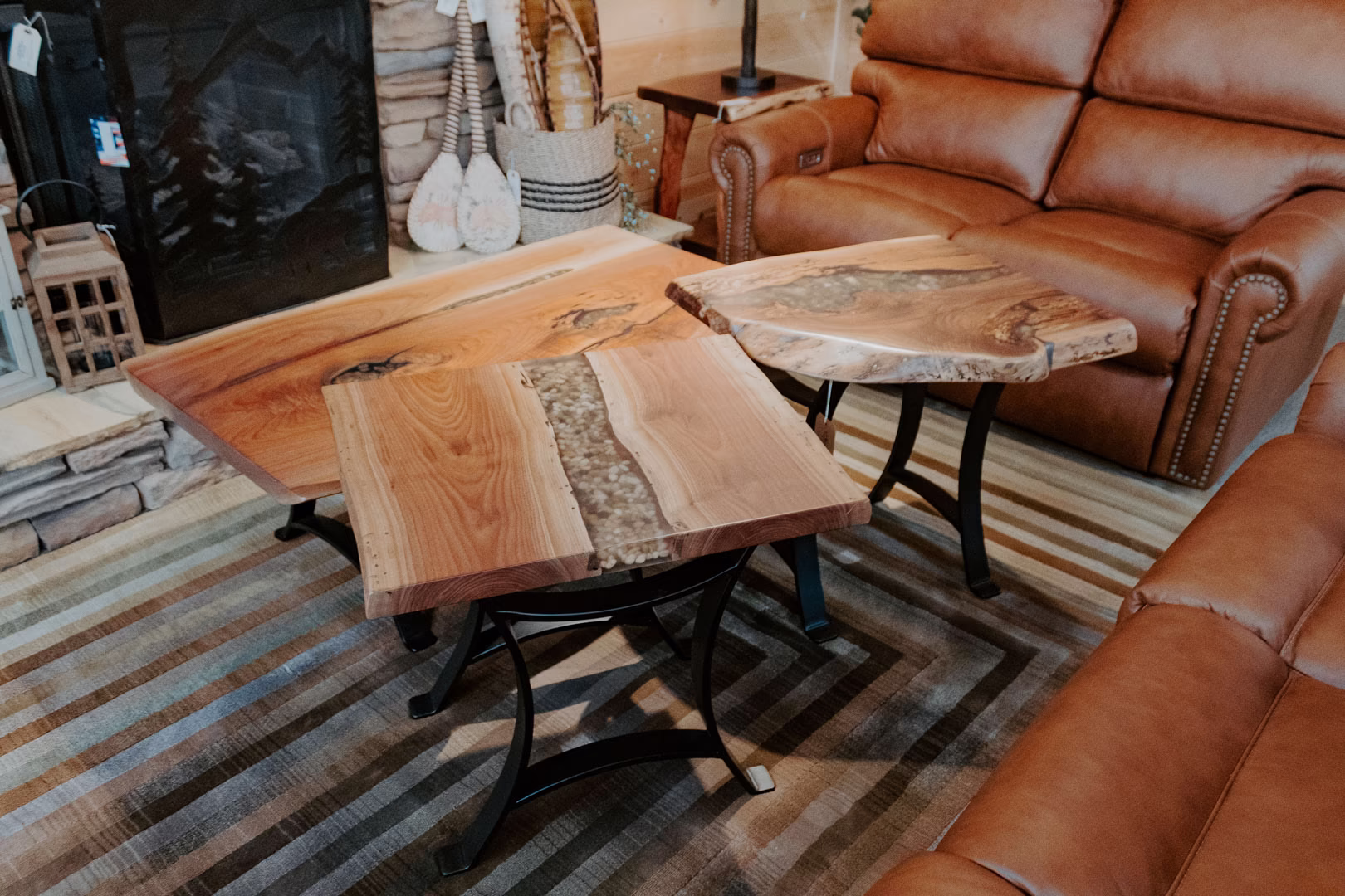 Three wooden coffee tables with natural wood finishes and metal legs are arranged in a living room, accompanied by a brown leather armchair, striped rug, and a fireplace.