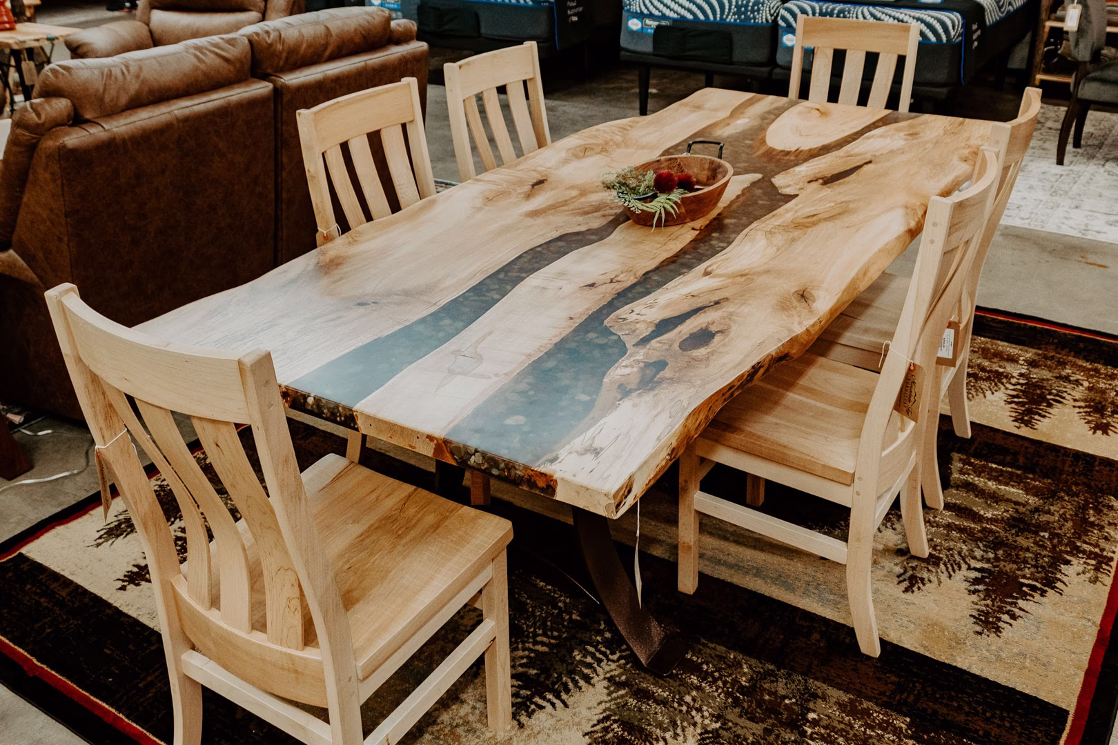 river-rock-maple-slab-collection-featured-image A light-colored wooden table with a black river stone top and six matching chairs sits in a furniture store, accompanied by a brown leather couch and a rug.