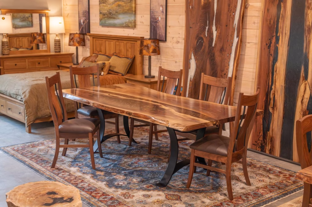 river-rock-walnut-slab-collection-featured-image A wooden dining table with a natural edge and four matching chairs sits on a blue and beige rug, accompanied by a wooden chest of drawers and paintings on a rustic wooden wall.