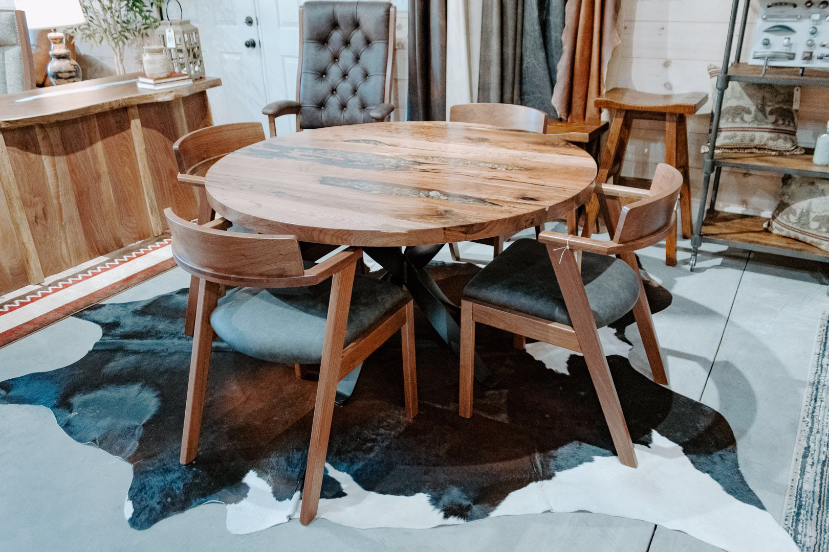 round-river-rock-walnut-collection-featured-image A round wooden table with six wooden chairs sits on a cowhide rug in a rustic store, surrounded by wooden furniture and decor.
