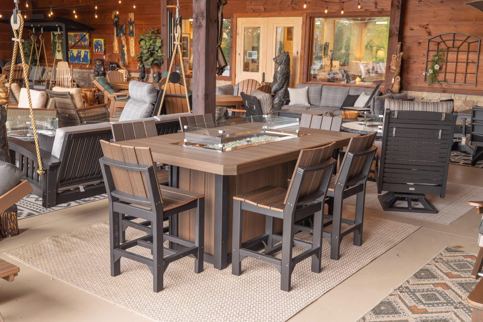A wooden patio features a table with a glass top, surrounded by black chairs, a gray sofa, a wooden swing, and plants, with a wooden archway in the background.