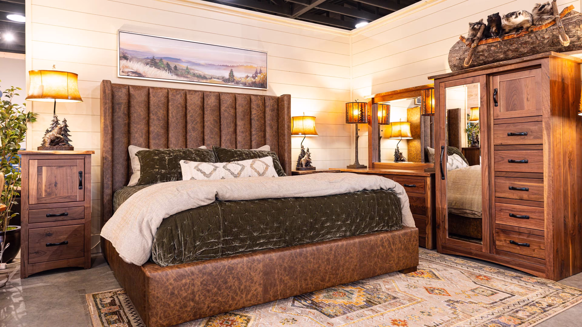 A luxurious bedroom features a brown leather bed with a white and gray comforter, flanked by wooden nightstands and lamps, with a large mirror and landscape painting on the white-painted walls.
