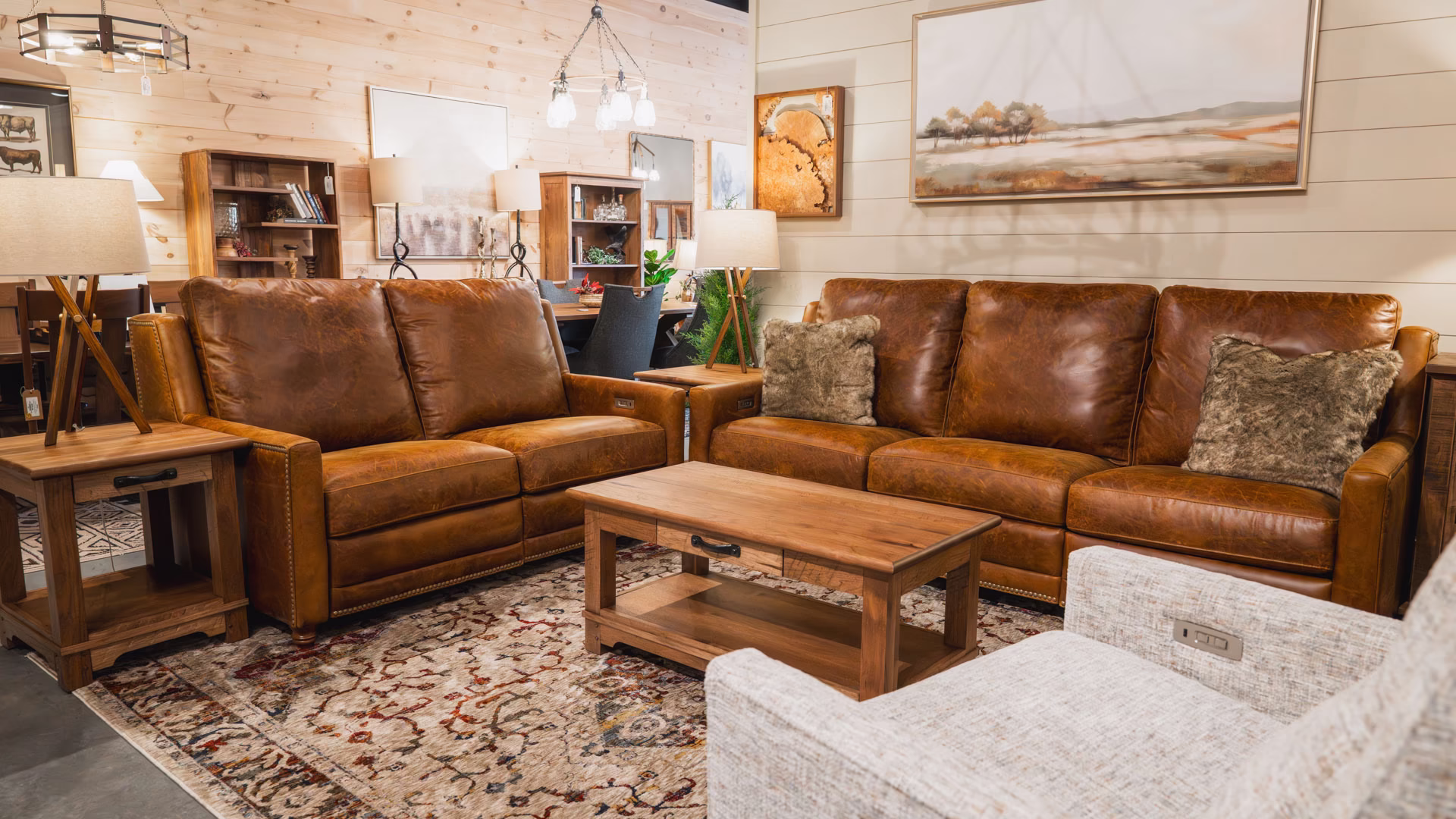 A brown leather sofa and loveseat with gray throw pillows, accompanied by a wooden coffee table and side table, fill a serene living room with wooden floors, walls, and a large abstract painting.
