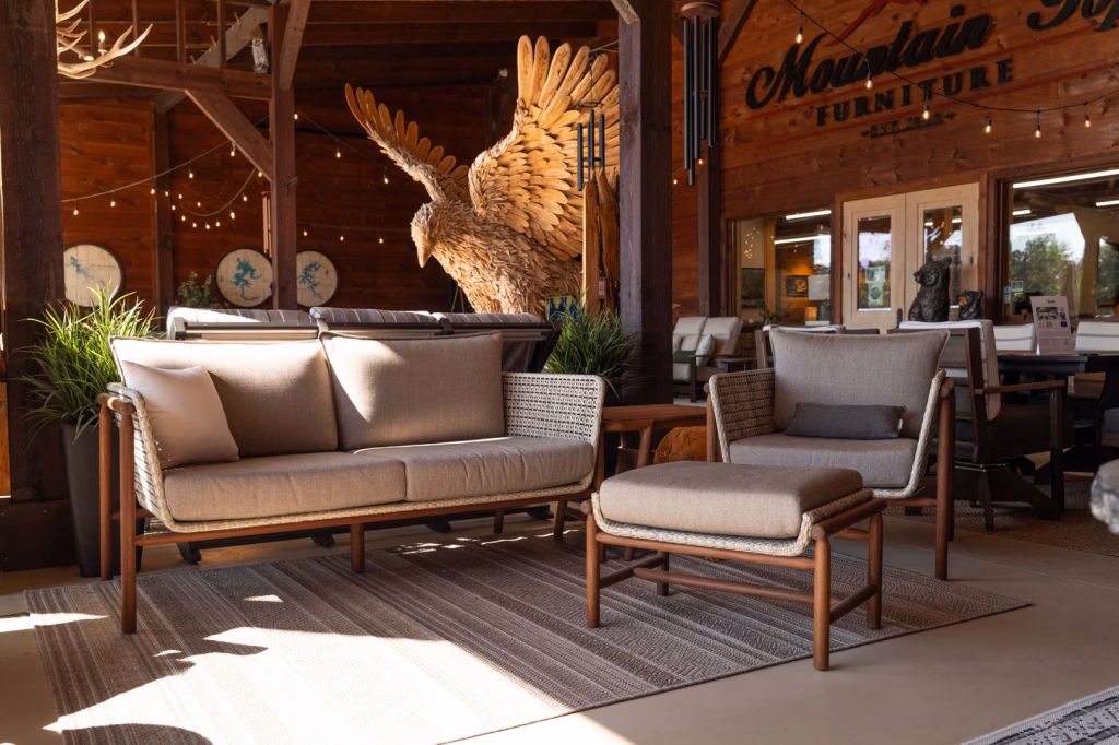 A modern beige outdoor furniture set, featuring a sofa, armchair, and ottoman on a striped rug, displayed in a rustic wood-paneled store with a large carved wooden eagle sculpture in the background.