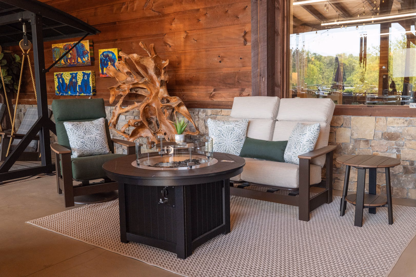 leeward-collection A cozy, rustic outdoor living space featuring a round fire pit table, a green armchair, and a cream loveseat arranged on a neutral rug against a wood-paneled wall.