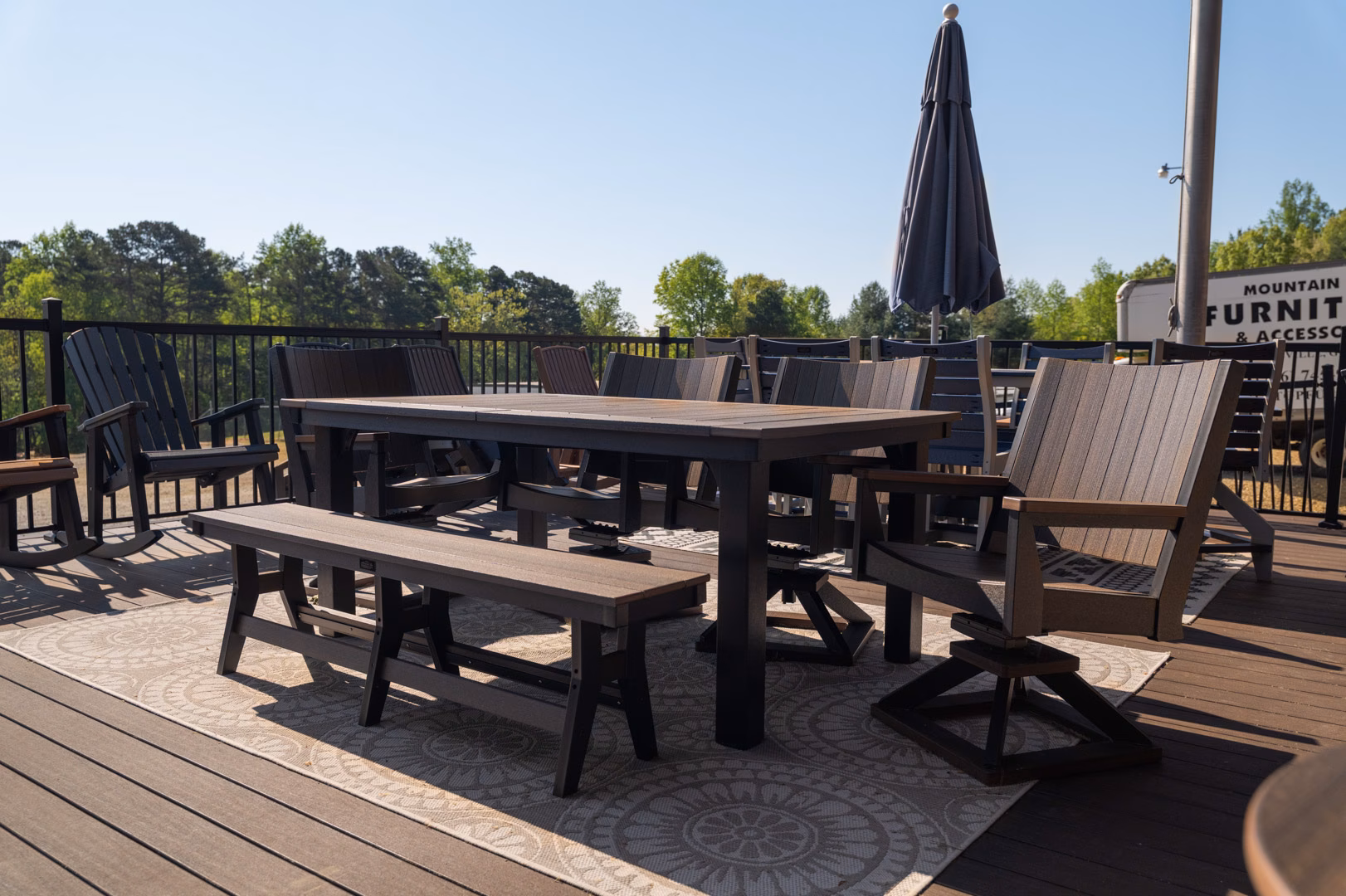 A brown outdoor dining set consisting of a table, bench, and several Adirondack chairs arranged on a patterned rug on a wooden deck.