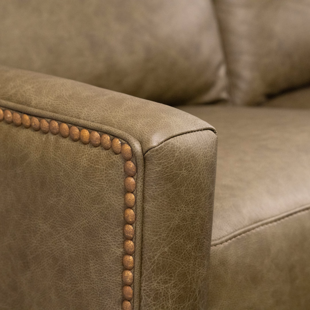 A close-up of the arm of a textured, olive-tan leather sofa featuring decorative nailhead trim along the seam.